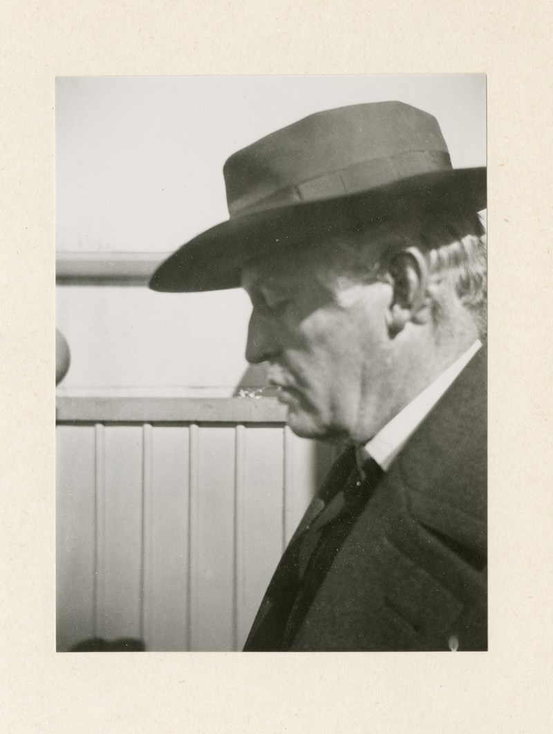A black and white photograph of Edvard Munch with his face in profile. He wears a suit jacket and a dark hat that casts a shadow over his closed eyes.