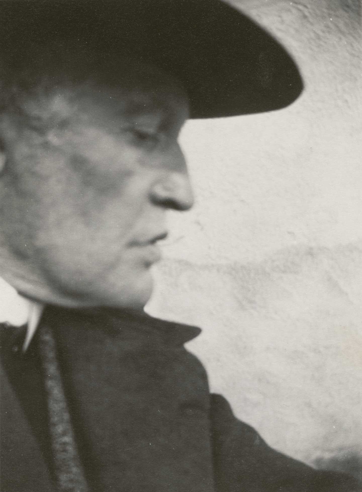 Et svart-hvitt fotografi av Edvard Munchs ansikt i profil. Han har på seg dressjakke, og en mørk hatt som kaster skygge over øynene