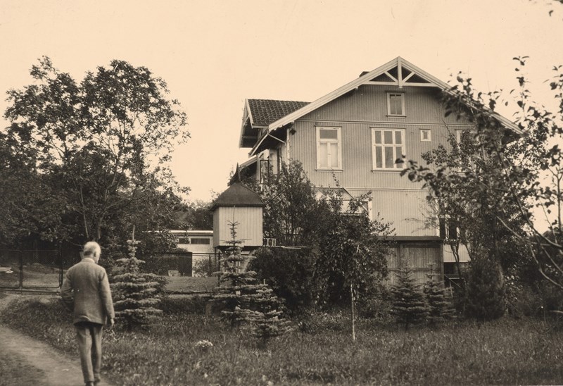Edvard Munch vandrer langs veien oppover mot Ekely. Det er en gresseng og trær foran huset.
