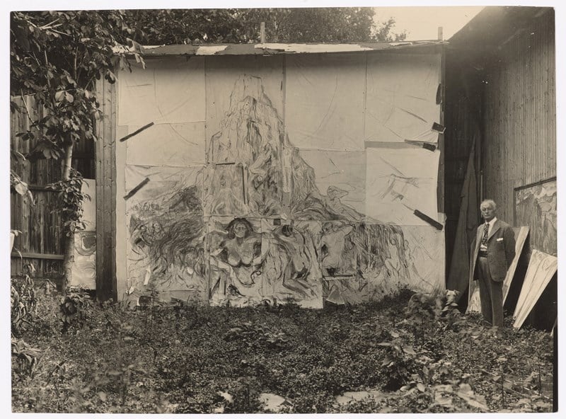 Munch’s open-air studio at Ekely. Edvard Munch is seen on the right side of the image, standing in front of the painting The Human Mountain. Photograph.
