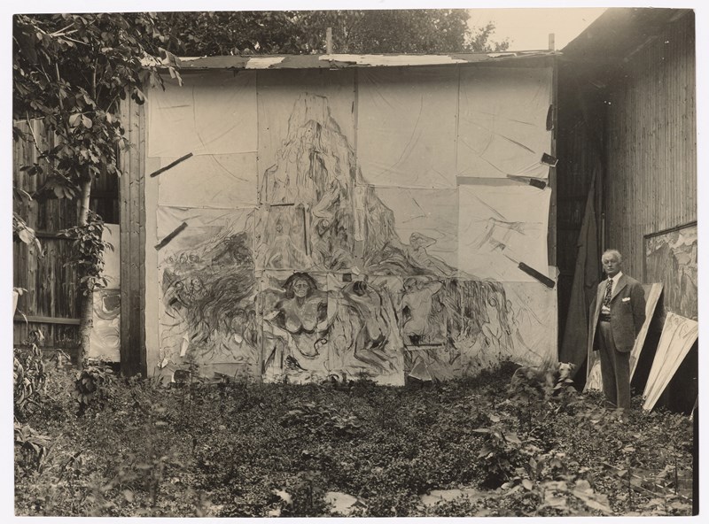 Munchs friluftsatelier på Ekely. Vi ser Edvard Munch til høyre i bildet. Han står foran maleriet Menneskeberget. Fotografi