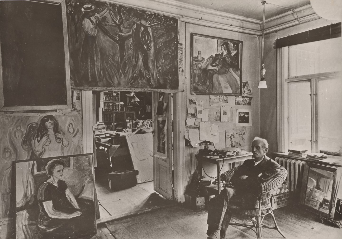 Edvard Munch sitter i en kurvstol i et rom på Ekely omgitt av sine malerier. Fotografi