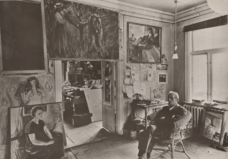 Edvard Munch sitter i en kurvstol i et rom på Ekely omgitt av sine malerier. Fotografi