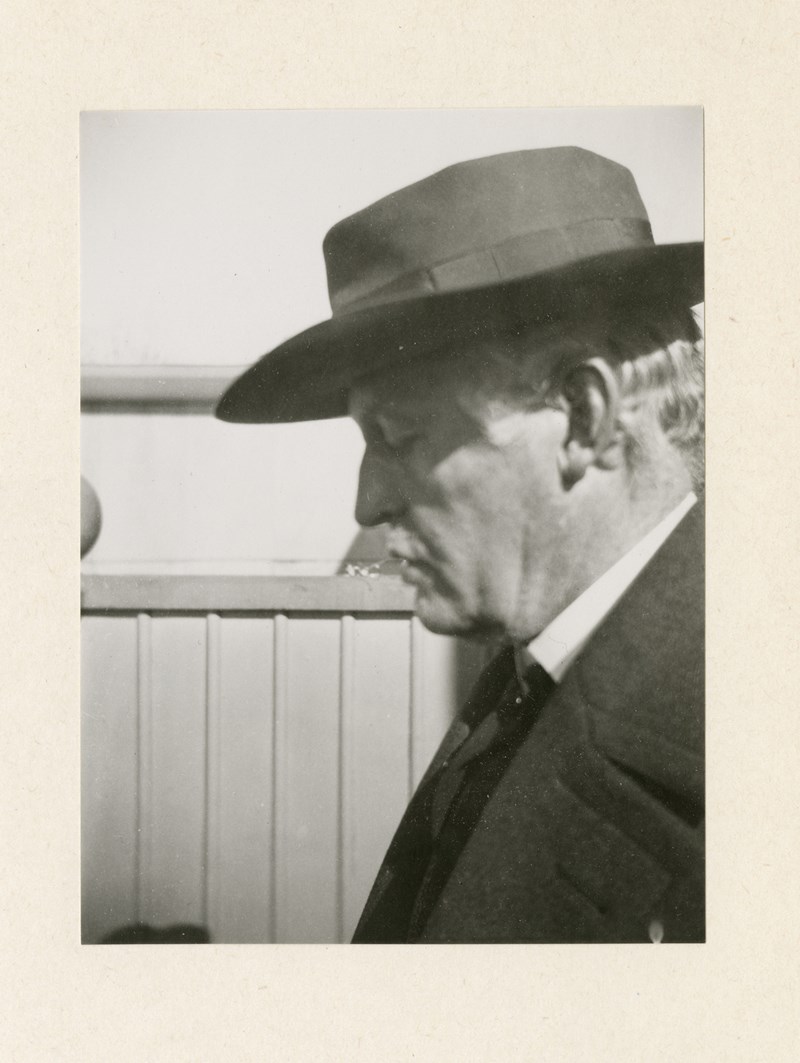 Et svart-hvitt fotografi av Edvard Munchs ansikt i profil. Han har på seg dressjakke, og en mørk hatt som kaster skygge over øynene, som er lukket. 