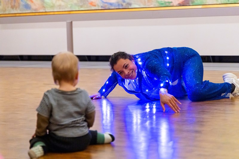 I forgrunnen, ute av fokus: en baby som sitter med ryggen til og iakttar en danser, i fokus, litt lengre bak i bildet, som delvis ligger dansende på gulvet og smiler mot barnet. Det blåglitrende kostymet til danseren gir gjenskinn i gulvet. 