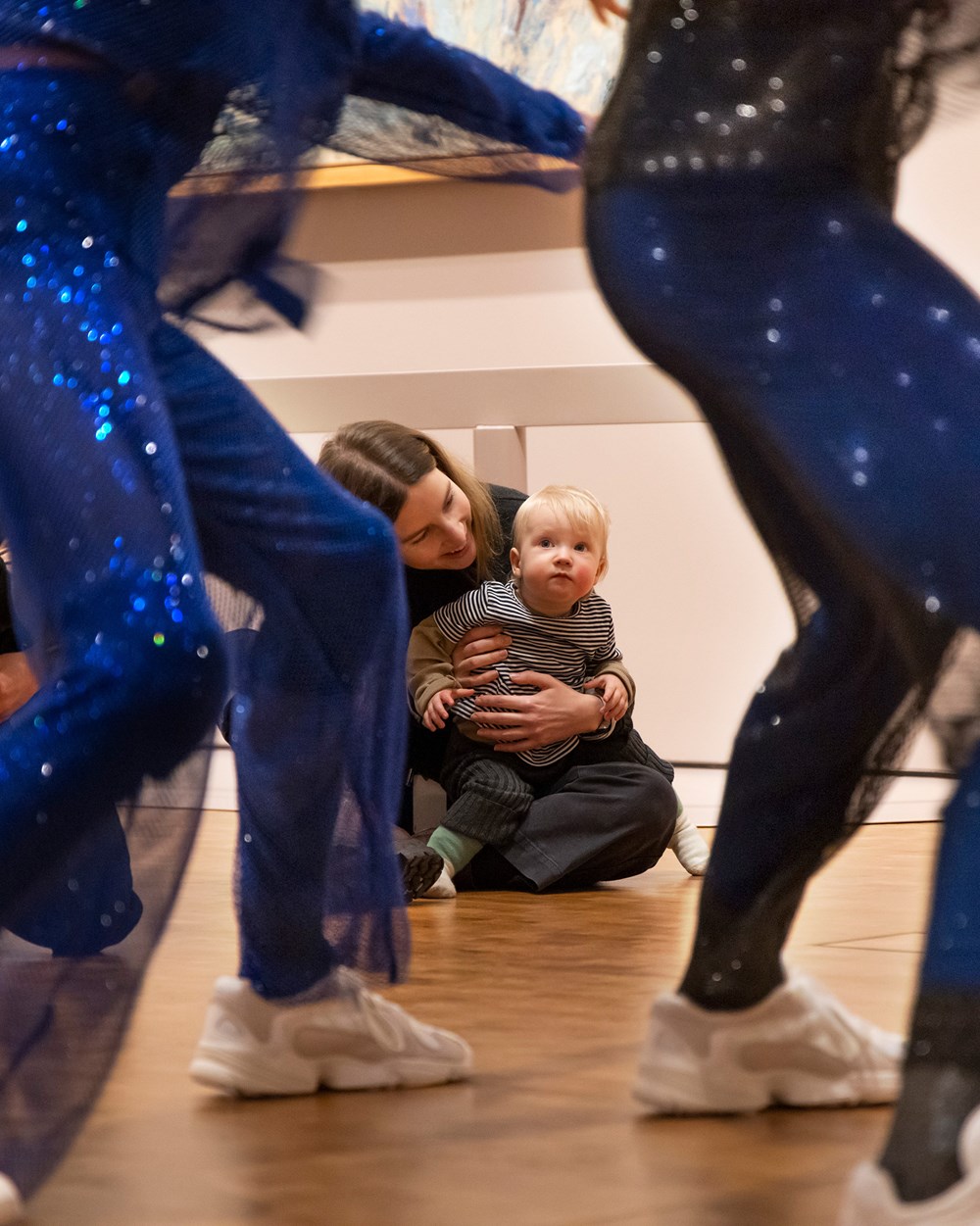I forgrunnen deler av kroppene til to dansere i blåglitrende kostymer, i bakgrunnen og i fokus en baby på mors fang som ser oppmerksomt på danserne