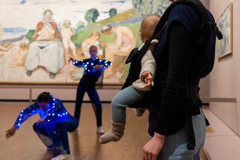 En baby i bæresele er vendt oppmerksomt mot to dansere i blåglitrende kostymer som danser foran Munchs monumentale verk