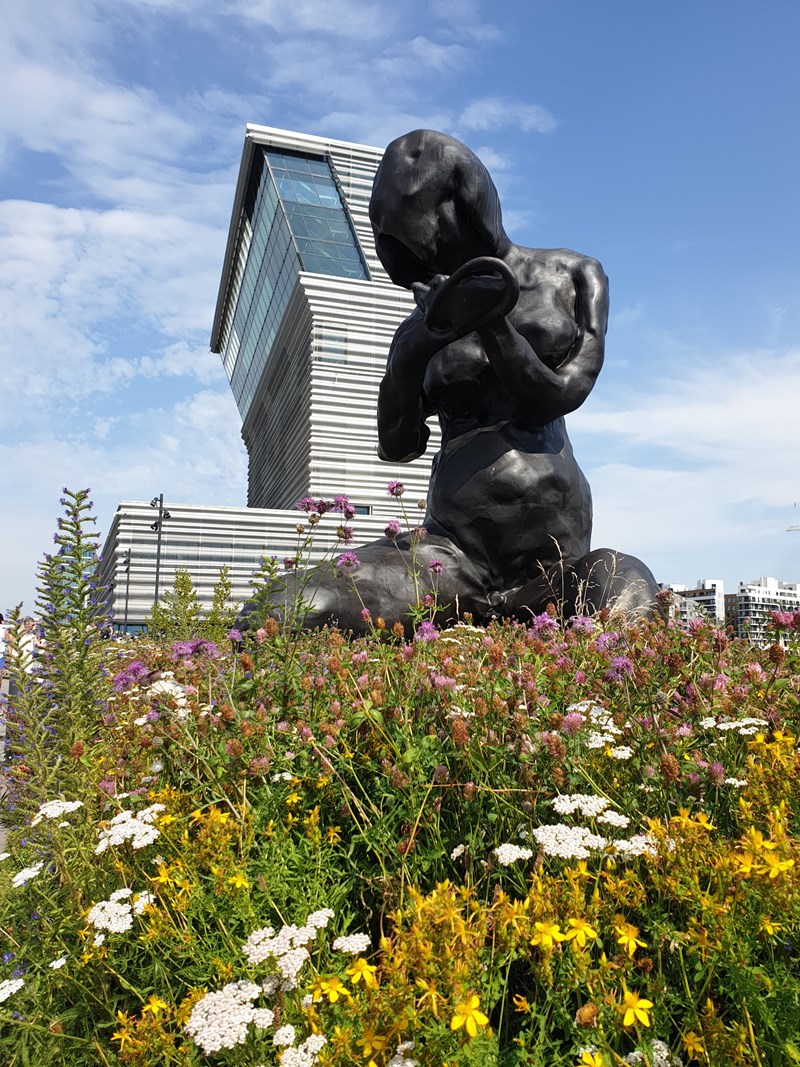 You see the sculpture The Mother, a dark bronze sculpture by artist Tracey Emin, standing 9 metres tall. In the background, you can see the MUNCH building and a blue sky. Photograph.