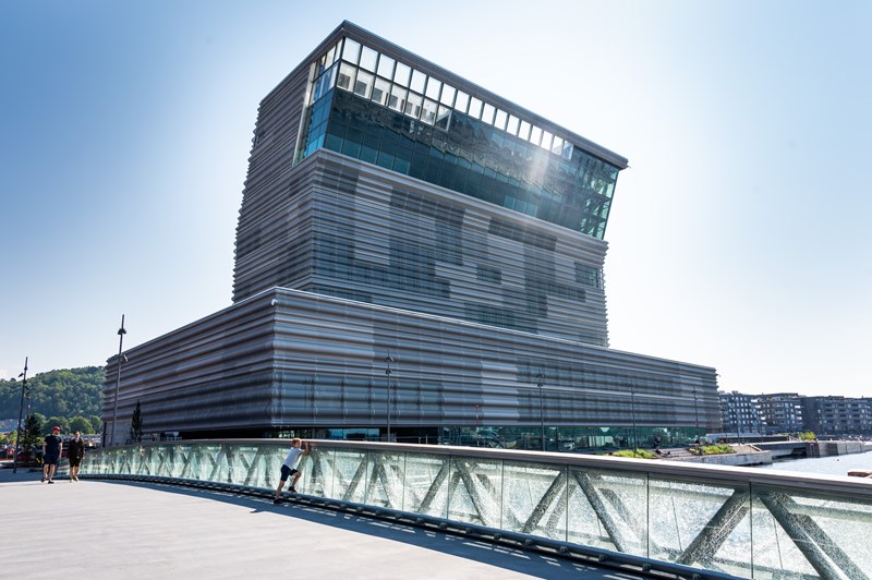 The new MUNCH in Bjørvika, seen from the west in a low-angle view. The sun is shining, and the sky is blue. In the foreground, you can see the bridge crossing the Akerselva River in front of the museum. Photograph.