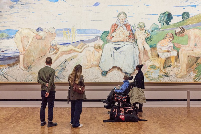 Four people stand with their backs to the camera, observing the painting Alma Mater by Edvard Munch. The artwork is 50 square metres in size. Photograph.