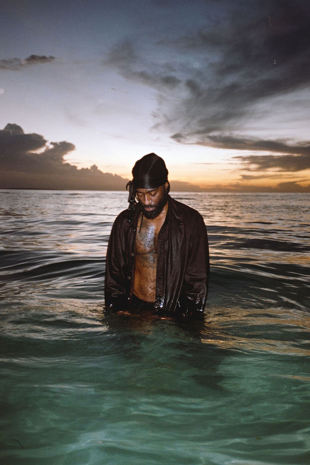 Arif står i havet med vann til skrittet og ser ned i vannet. Han har en svart skjorte som er åpen og det er solnedgang i bakgrunnen. Foto