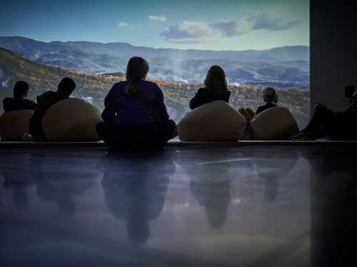 Installasjonsbilde fra Corpus Infinitum. Publikum sitter på puter på gulvet foran store projiseringer av natur. 