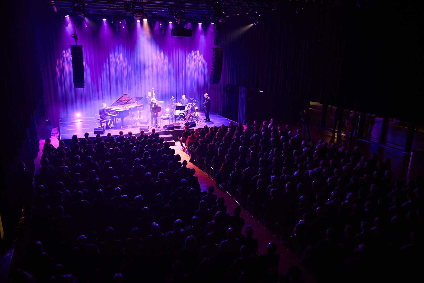 En fullsatt festsal omgitt av mørkt rosalilla lys, og spotlys mot scenen, hvor en gruppe jazzmusikere skimtes.