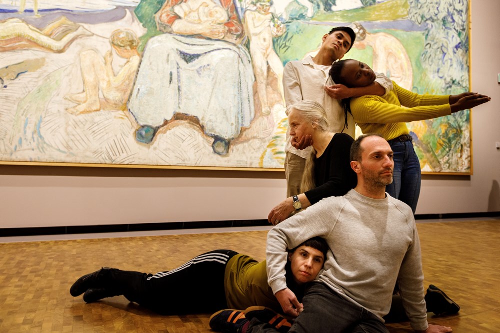 Five dancers in front of Edvard Munch's painting Alma Mater, performing Permanent Collection. They stand, sit, and lie intertwined with one another. Photograph.