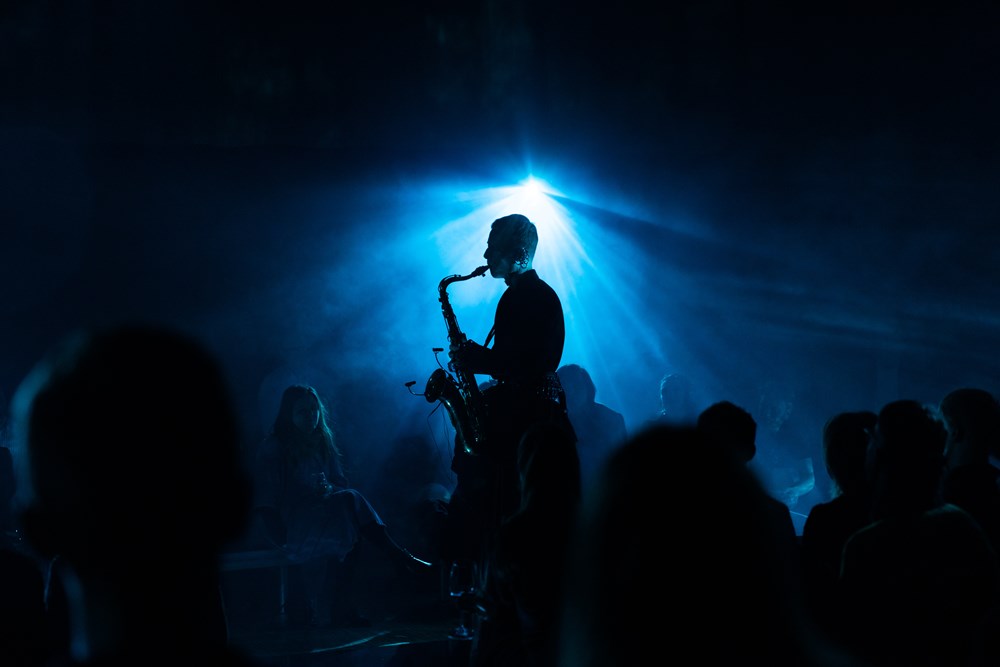 Saxophonist Bendik Giske stands and plays in the middle of a crowd seated on the floor, listening. The room is dark, illuminated by blue light. Photograph.