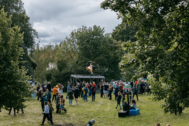 In the middle of the lush, green Tøyen Park, a tall stage has been set up where a man is dancing with flowing ribbons. A crowd stands on the grassy slope, watching him. Photograph.