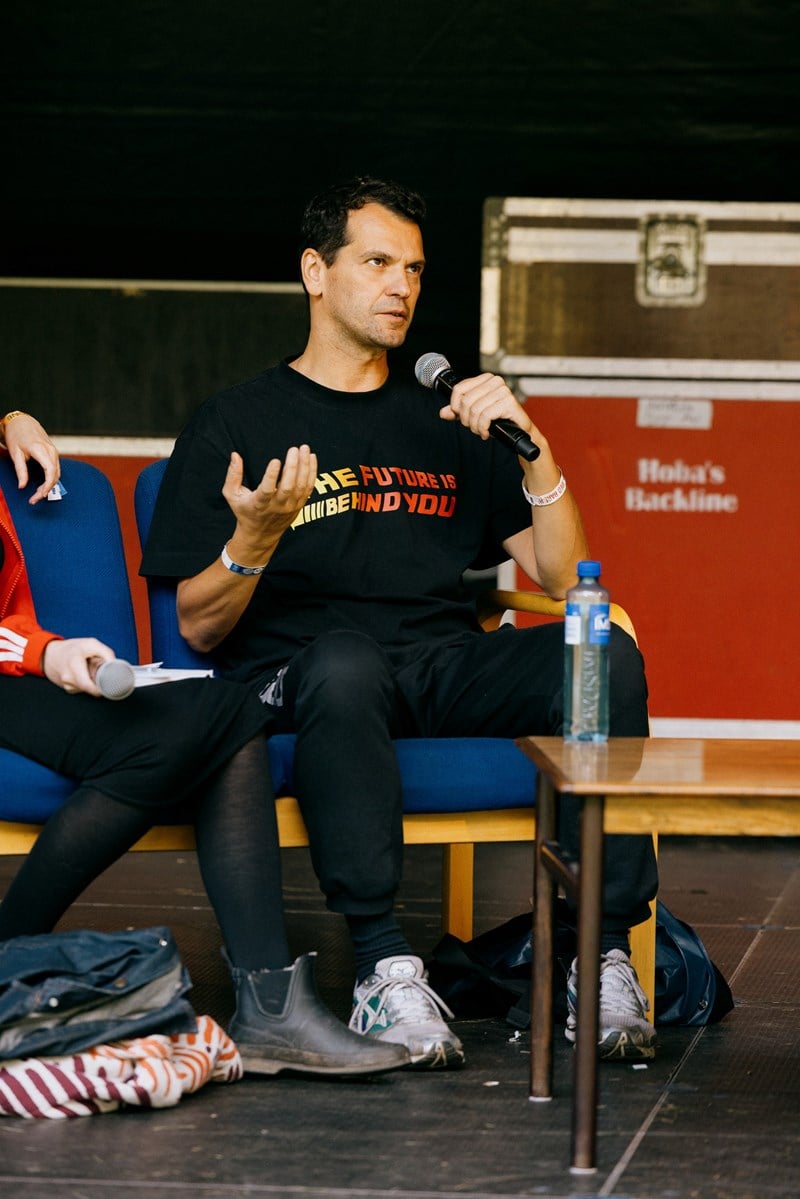 Performance artist Manuel Pelmuș sits on stage, speaking into a handheld microphone. Photograph.