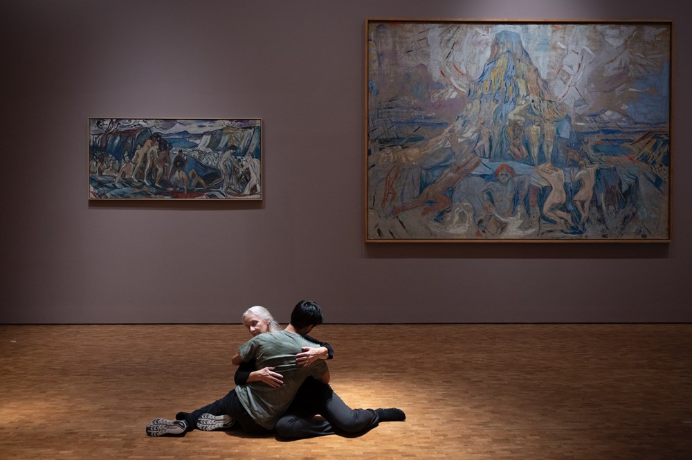 To mennesker sitter på gulvet i en omfavnelse. I bakgrunnen ser du maleriet Menneskeberget av Edvard Munch. Fotografi