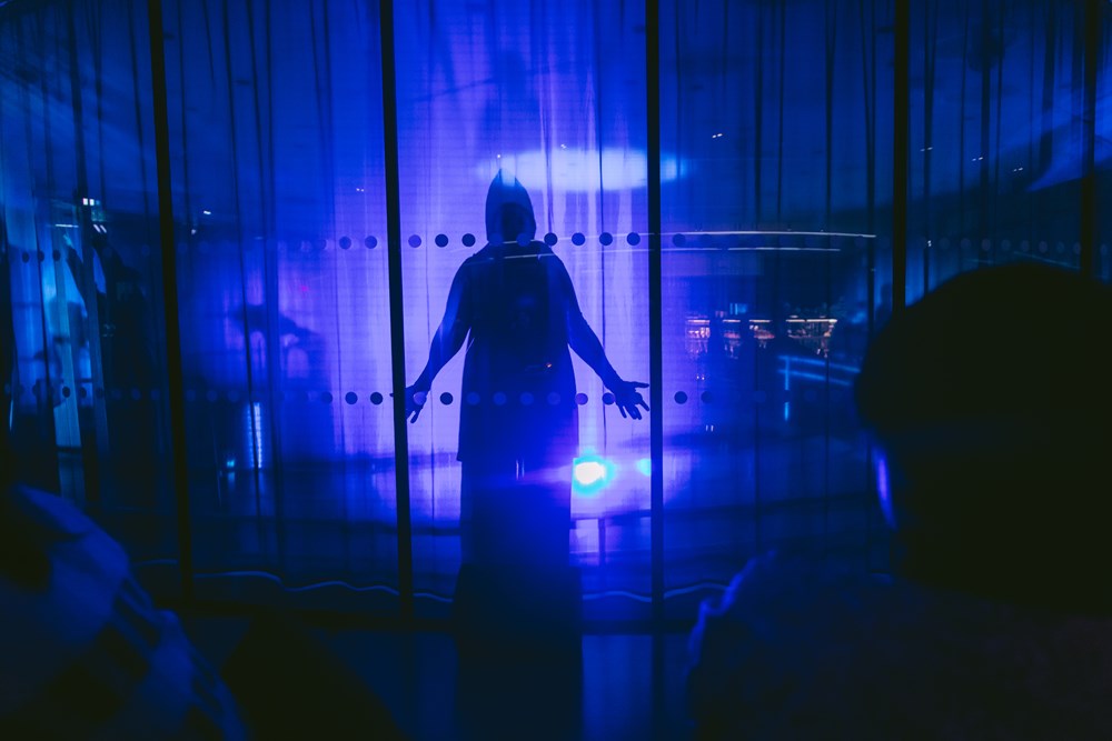 A cloaked figure stands behind a glass wall in a room illuminated by blue light. Photograph.